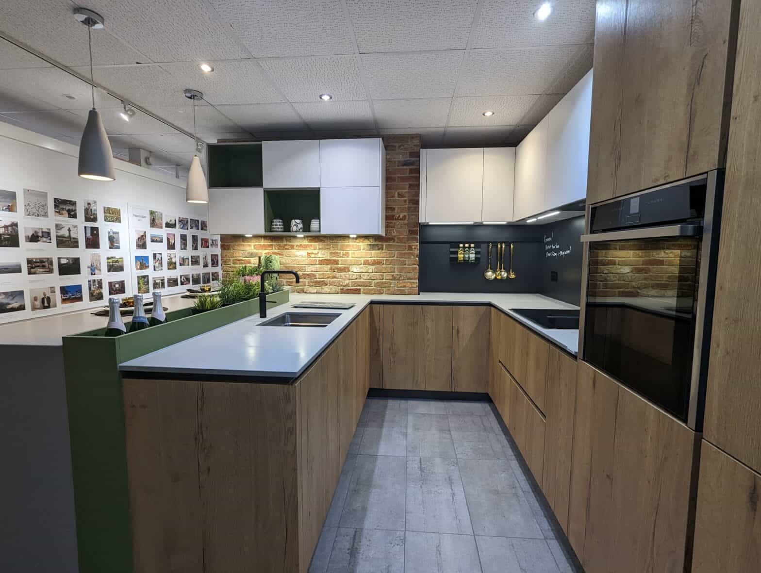 A photo of a U shaped kitchen with wood effect doors and concrete worktop. There is an oven in a tall housing. The splash backs are a writable chalk board like panel.