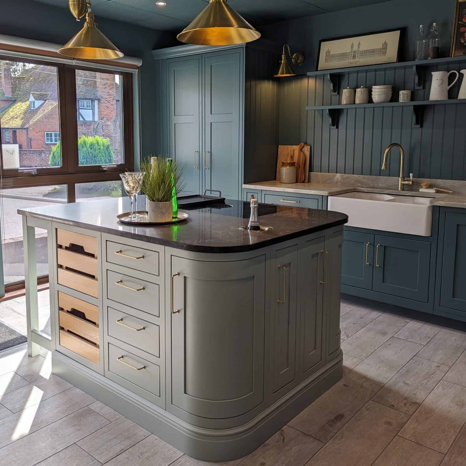 A photo of a traditional shaker style kitchen in blue with a blue/green painted island that has rounded edges to one side and a breakfast bar to the other. Tongue and groove panels feature on the back wall behind the sink with a granite worktop and upstand.