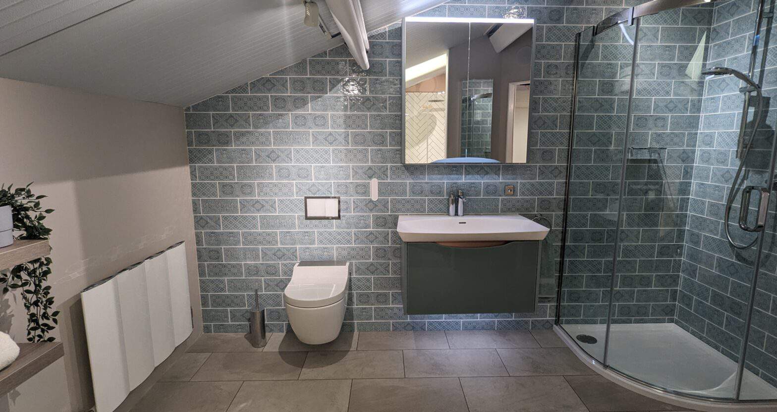 A photo of a bathroom display. Blue textured tiles span across the far wall and wrap into the corner shower enclosure on the right. A complimentary blue wash basin sits in the middle with a mirrored cabinet above and a wall hung toilet on the left with hidden cistern.