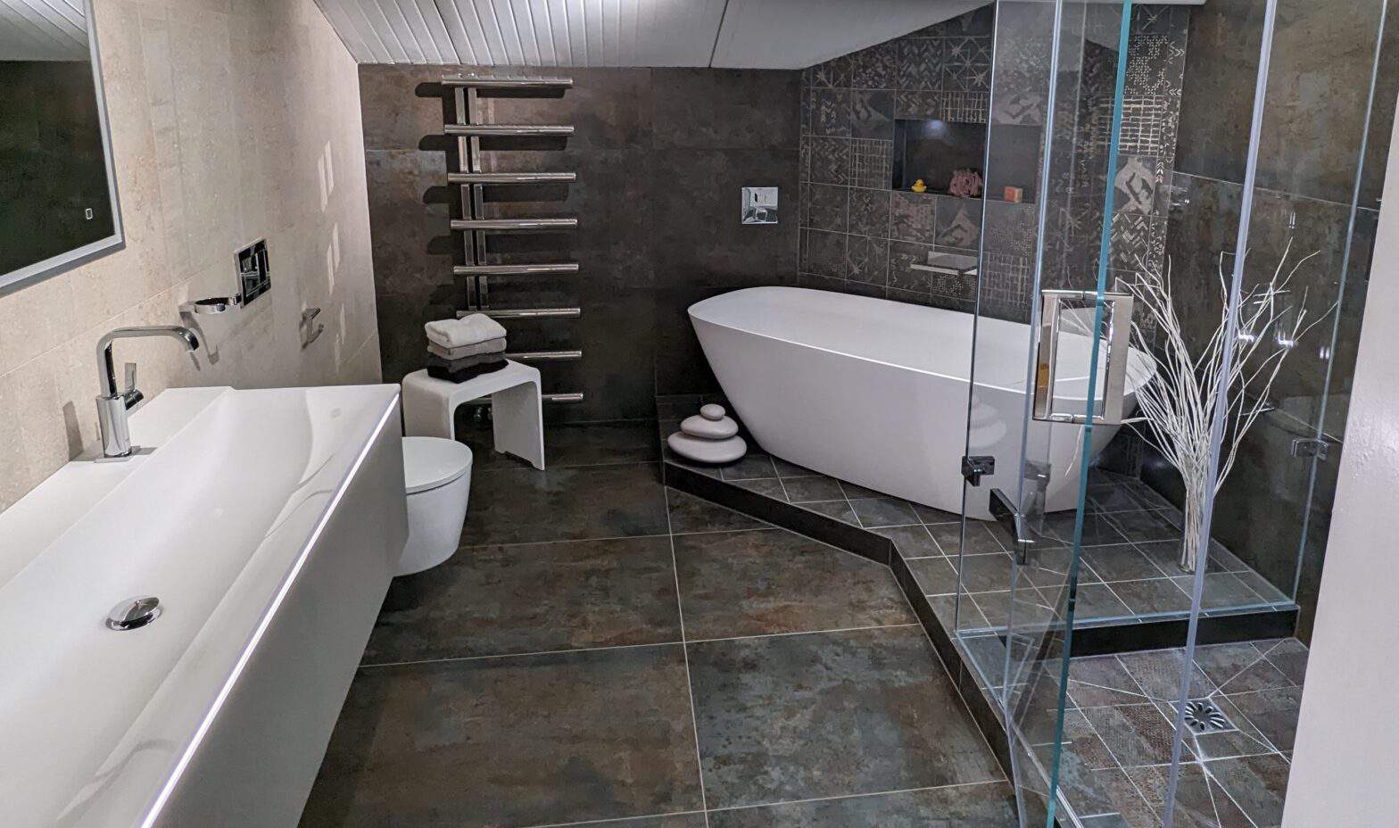 A photo of a luxury bathroom featuring a free standing bath on a raised platform that runs into a floor to ceiling shower/steam room. A double basin and vanity with LED mirror are on the left.