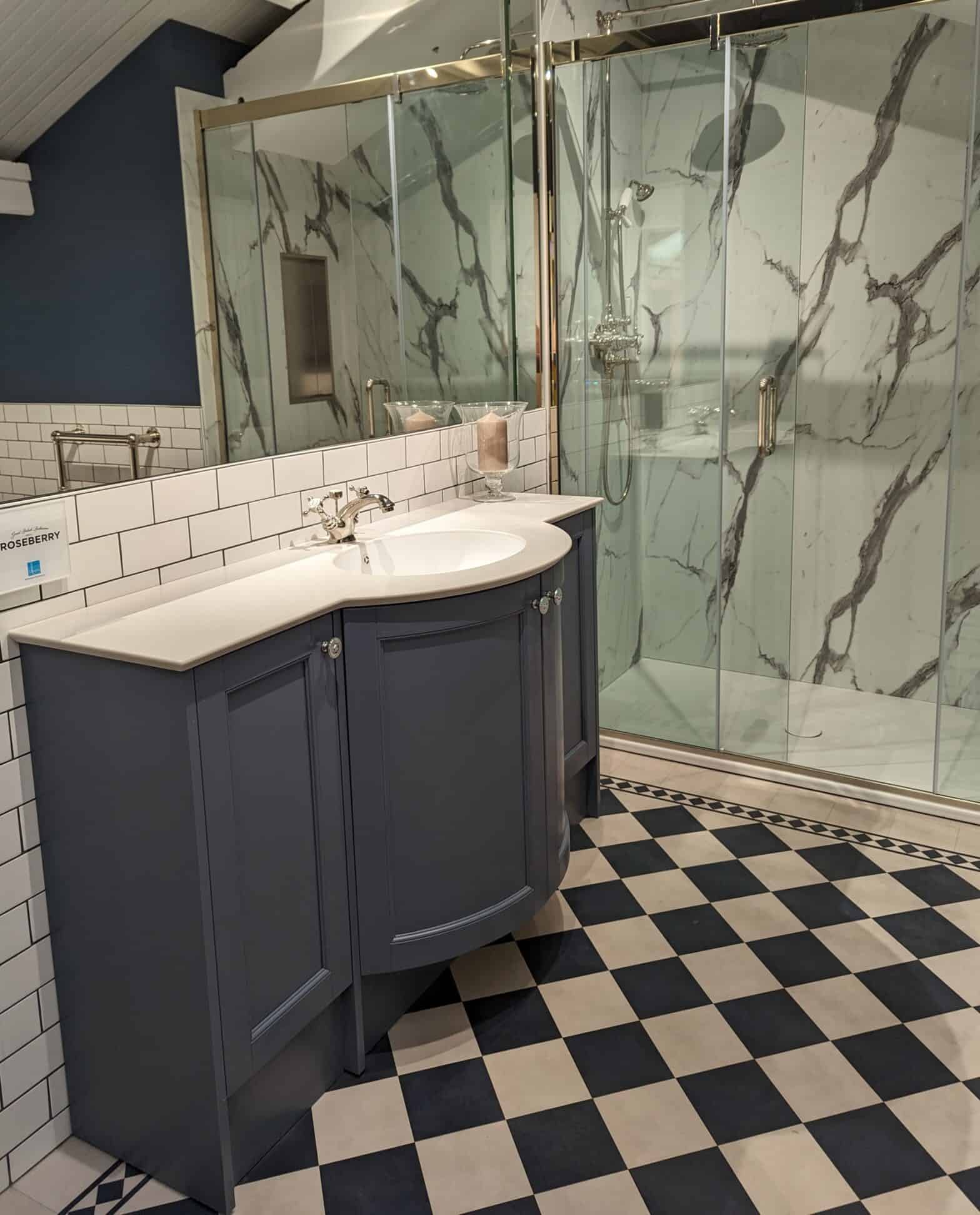 A photo of a traditional style bathroom with large walk in shower with sliding door and marble effect wall panels. A floor standing vanity unit sits along a wall tiled in white subway tiles.