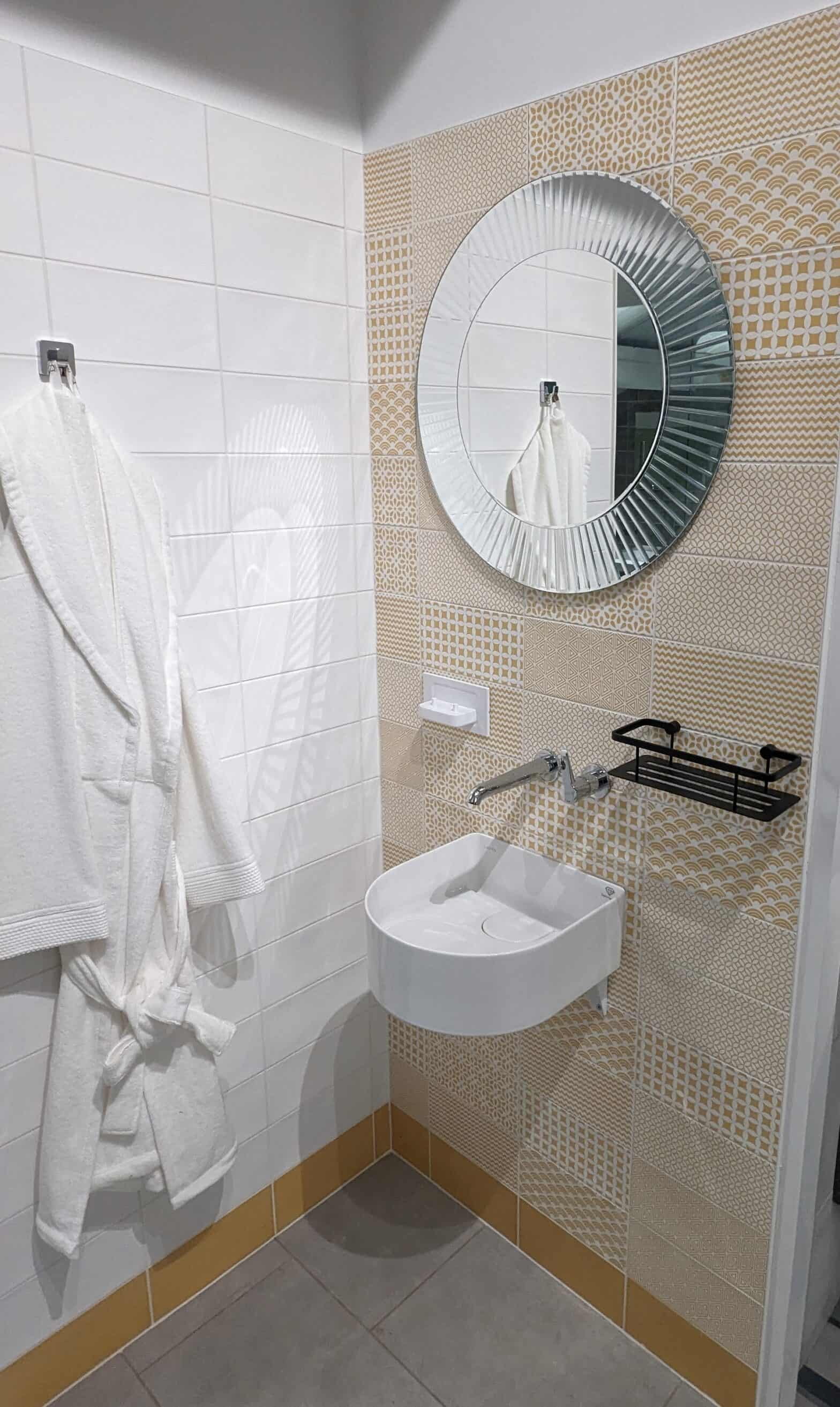 A photo of corner of a bathroom featuring white tiles on the left wall and yellow and white patterned tiles on the right with a wall hung basin and tap with wall hung lever.