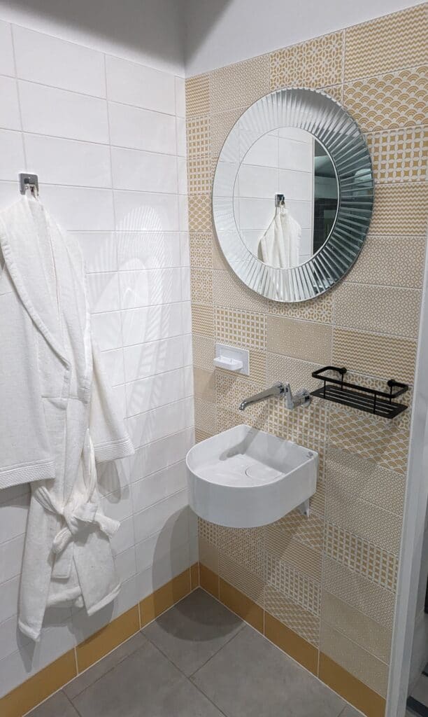 A photo of our corner display with yellow and white tiles, round mirror, white basin and black accents