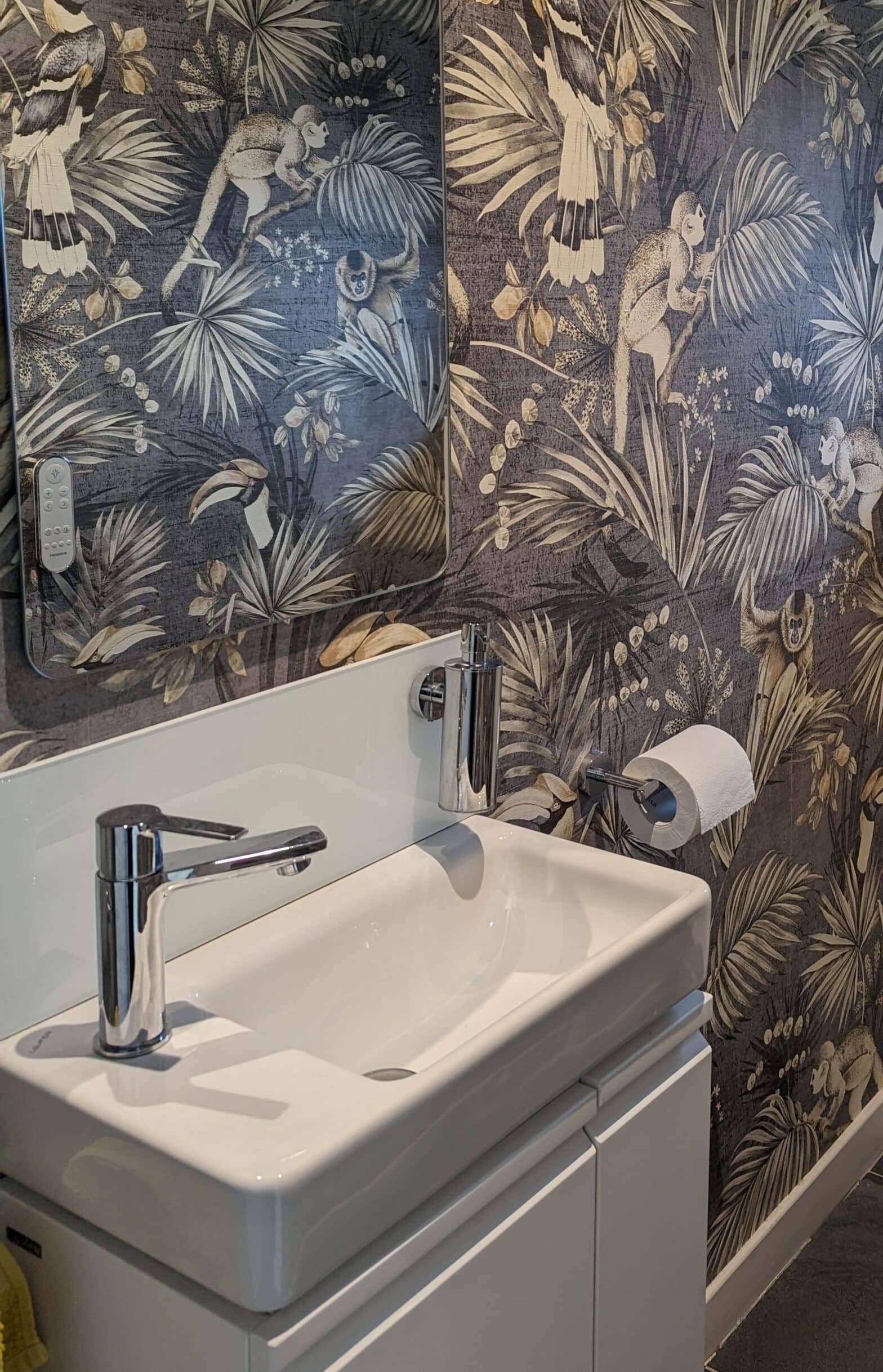A photo of a cloakroom featuring a tropical wallpaper with large scale leaves, birds and monkeys in a muted colour palette with slimline basin and vanity in white.